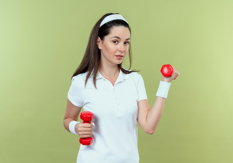Foto grátis mulher jovem fitness com uma faixa na cabeça, malhando com halteres, parecendo confiante em pé sobre a parede de luz