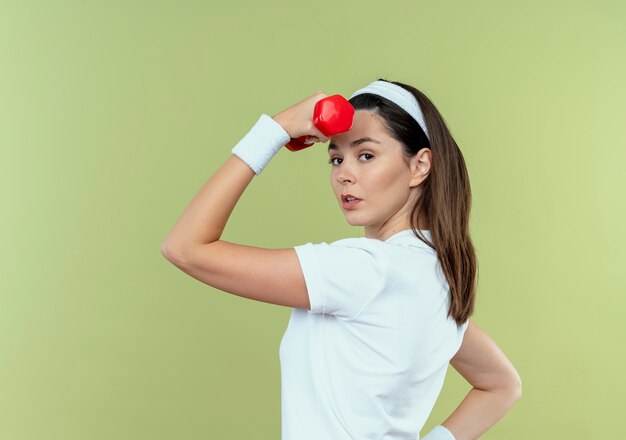 Mulher jovem fitness com uma faixa na cabeça, malhando com halteres, parecendo confiante em pé sobre um fundo claro