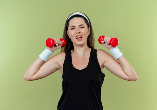 Mulher jovem fitness com uma faixa na cabeça, malhando com halteres, parecendo confusa em pé sobre um fundo claro