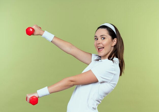 Mulher jovem fitness com uma faixa na cabeça malhando com halteres sorrindo alegremente feliz e positiva em pé sobre um fundo claro