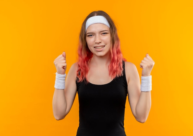 Mulher jovem fitness em roupas esportivas cerrando os punhos feliz e exultante, regozijando-se com seu sucesso em pé sobre a parede laranja
