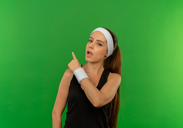 Mulher jovem fitness em roupas esportivas com bandana confusa apontando com o dedo para o lado em pé sobre a parede verde