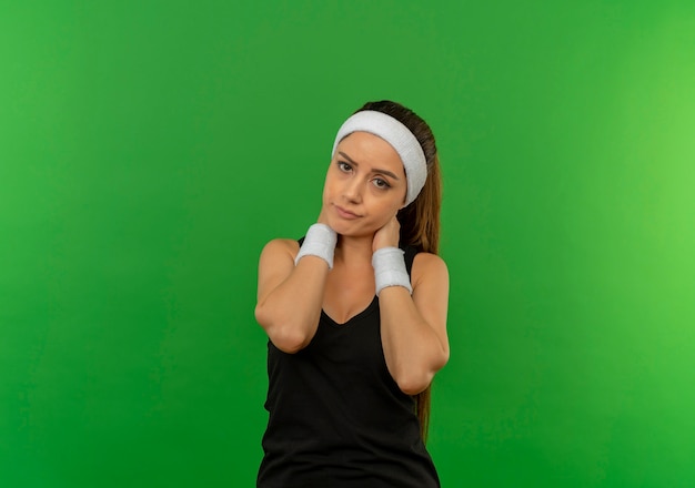 Mulher jovem fitness em roupas esportivas com bandana parecendo doente, tocando seu pescoço, com dor em pé sobre a parede verde