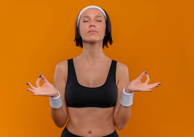 Foto grátis mulher jovem fitness em roupas esportivas com bandana relaxando com os olhos fechados fazendo gestos de meditação com os dedos em pé sobre a parede laranja