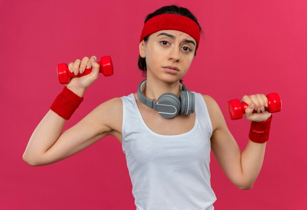 Mulher jovem fitness em roupas esportivas com bandana segurando dois halteres fazendo exercícios com expressão triste no rosto em pé sobre a parede rosa