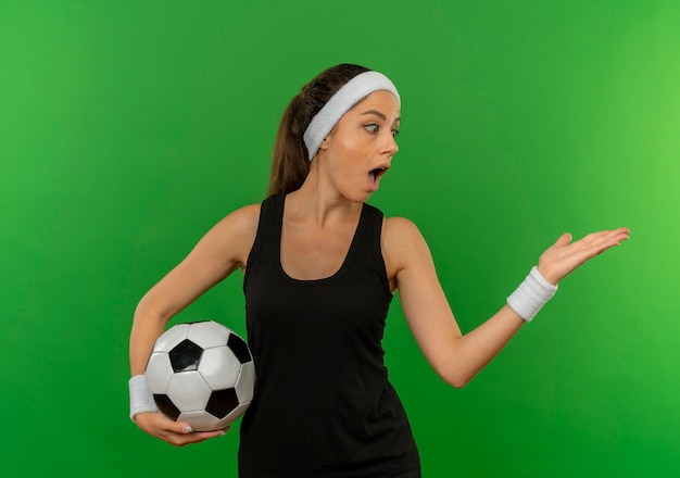 Mulher jovem fitness em roupas esportivas com bandana segurando uma bola de futebol apontando com o braço da mão para o lado, parecendo surpresa em pé sobre a parede verde