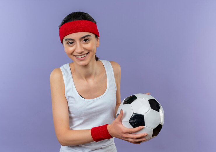Foto grátis mulher jovem fitness em roupas esportivas com bandana segurando uma bola de futebol com um sorriso no rosto em pé sobre a parede roxa