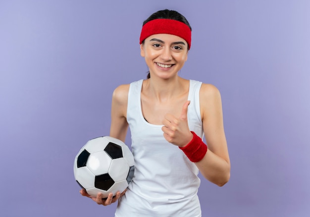 Mulher jovem fitness em roupas esportivas com bandana segurando uma bola de futebol, sorrindo confiante mostrando os polegares em pé sobre a parede roxa