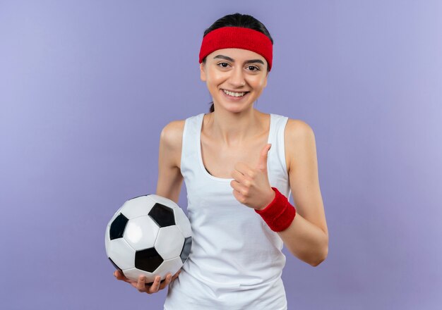 Mulher jovem fitness em roupas esportivas com bandana segurando uma bola de futebol, sorrindo confiante mostrando os polegares em pé sobre a parede roxa