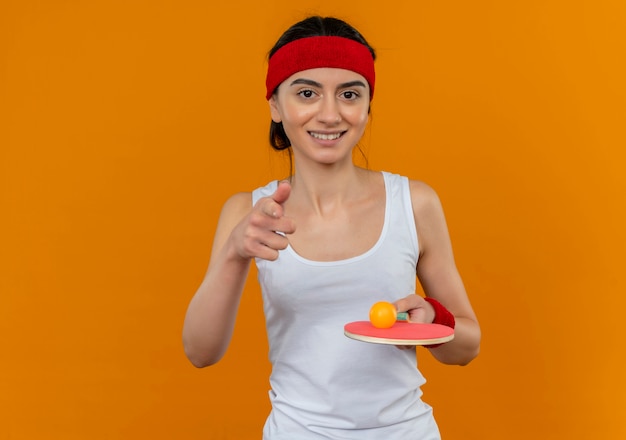 Mulher jovem fitness em roupas esportivas com bandana segurando uma raquete e uma bola de tênis de mesa sorrindo com uma cara feliz