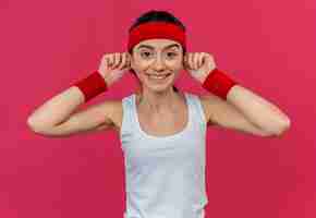 Foto grátis mulher jovem fitness em roupas esportivas com bandana tocando suas orelhas com um grande sorriso no rosto em pé sobre a parede rosa
