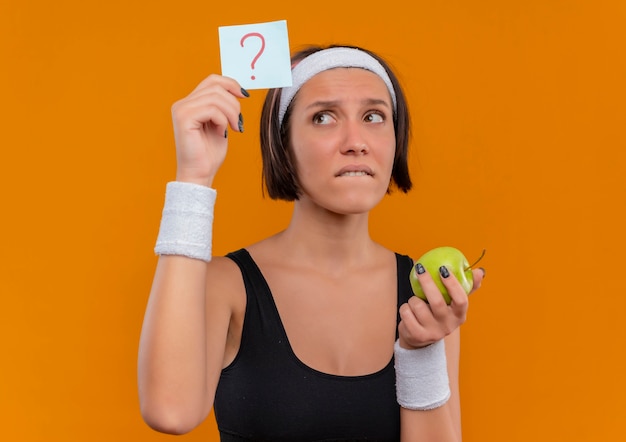 Mulher jovem fitness em roupas esportivas com fita para a cabeça mostrando papel lembrete com ponto de interrogação segurando uma maçã verde olhando para o papel confuso em pé sobre a parede laranja