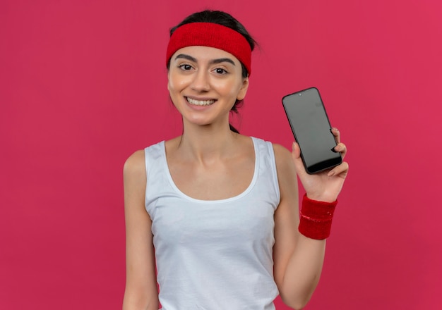 Mulher jovem fitness em roupas esportivas com fita para a cabeça mostrando smartphone feliz e positivo sorrindo alegremente em pé sobre a parede rosa