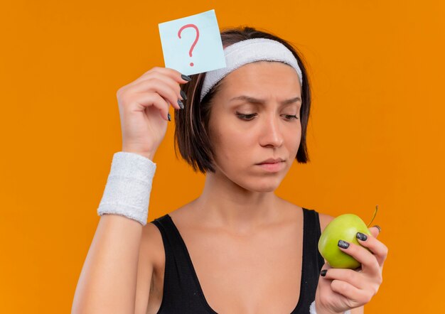 Mulher jovem fitness em roupas esportivas com fita para a cabeça segurando papel lembrete com ponto de interrogação e maçã verde olhando para uma maçã com expressão pensativa em pé sobre a parede laranja