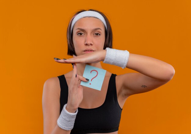 Mulher jovem fitness em roupas esportivas com fita para a cabeça segurando um papel lembrete com ponto de interrogação fazendo gesto de tempo com as mãos com cara séria em pé sobre a parede laranja