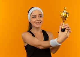 Foto grátis mulher jovem fitness em roupas esportivas com fita para a cabeça segurando um troféu feliz e positiva sorrindo alegremente em pé sobre a parede laranja