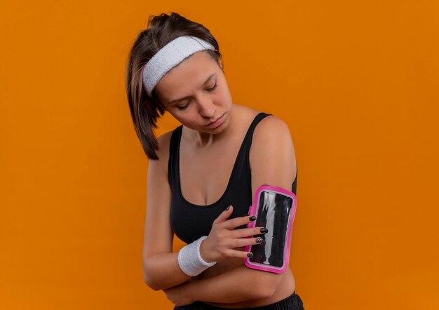 Mulher jovem fitness em roupas esportivas com fita para a cabeça tocando a braçadeira do smartphone em pé sobre a parede laranja