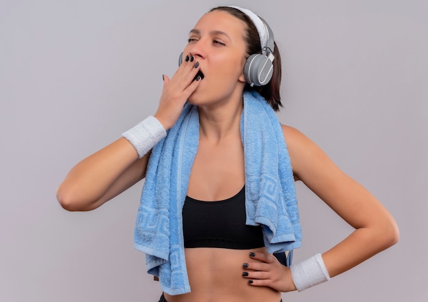 Mulher jovem fitness em roupas esportivas com fones de ouvido na cabeça e uma toalha no pescoço cansada quer dormir bocejando em pé sobre uma parede branca