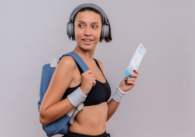 Mulher jovem fitness em roupas esportivas com fones de ouvido na cabeça segurando a passagem aérea olhando para o lado com um sorriso no rosto em pé sobre uma parede branca