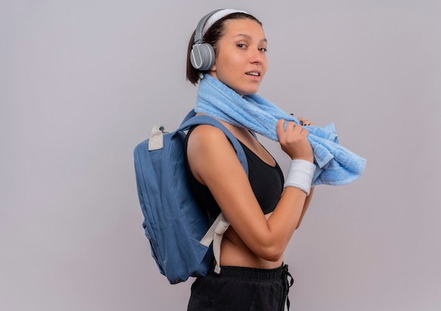 Mulher jovem fitness em roupas esportivas com mochila e fones de ouvido na cabeça com uma toalha no pescoço parecendo confiante sorrindo em pé sobre uma parede branca