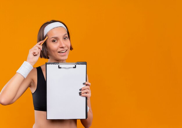 Mulher jovem fitness em roupas esportivas com tiara mostrando a prancheta com páginas em branco apontando com uma caneta para sua têmpora, parecendo confiante em pé sobre a parede laranja