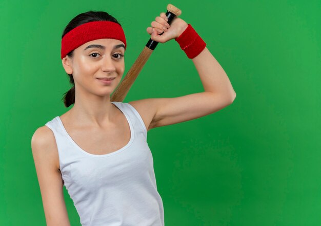 Foto grátis mulher jovem fitness em roupas esportivas com tiara segurando o taco de beisebol com um sorriso confiante em pé sobre a parede verde