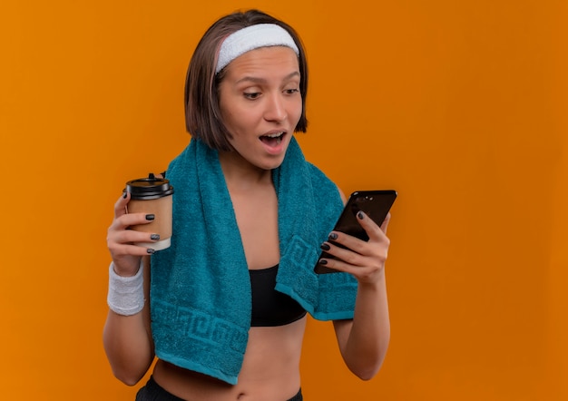 Mulher jovem fitness em roupas esportivas com uma toalha no pescoço segurando a xícara de café, olhando para a tela de seu smartphone surpresa e feliz em pé sobre a parede laranja