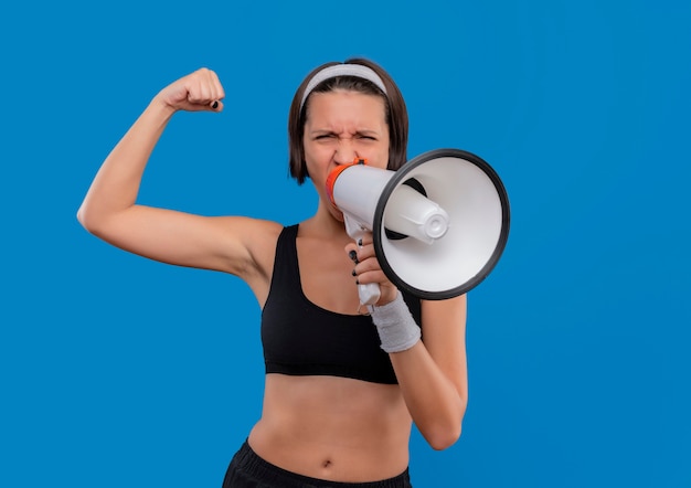 Mulher jovem fitness em roupas esportivas gritando para o megafone com expressão agressiva levantando o punho, conceito vencedor em pé sobre a parede azul