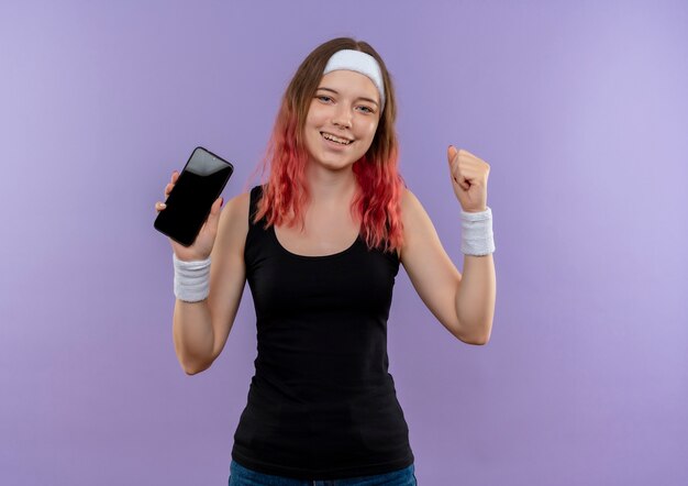 Mulher jovem fitness em roupas esportivas mostrando o punho cerrado de smartphone sorrindo alegremente feliz e saindo em pé sobre a parede roxa