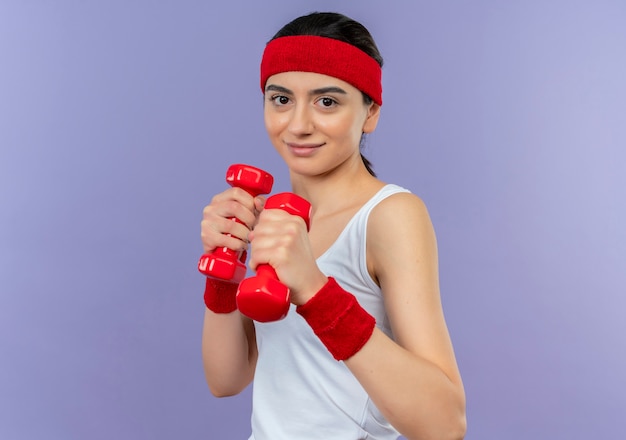 Mulher jovem fitness em roupas esportivas segurando dois halteres, posando como um boxeador, parecendo confiante em pé sobre a parede roxa