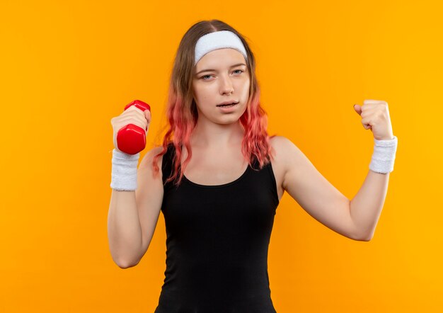 Mulher jovem fitness em roupas esportivas segurando halteres levantando o punho como uma vencedora em pé sobre uma parede laranja