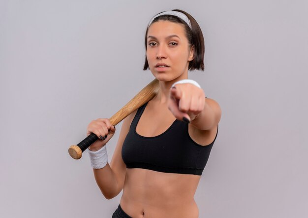 Mulher jovem fitness em roupas esportivas segurando taco de beisebol apontando com o dedo para você, parecendo confiante em pé sobre uma parede branca