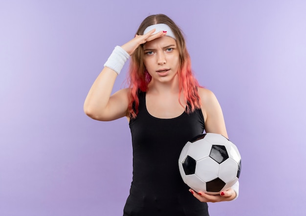 Mulher jovem fitness em roupas esportivas segurando uma bola de futebol com a mão na cabeça em pé sobre a parede roxa