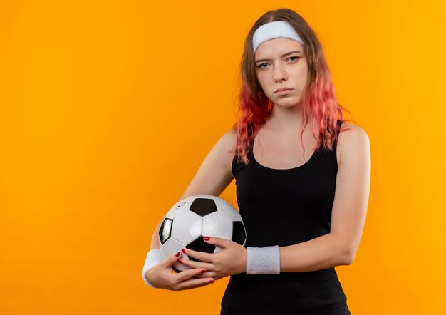 Mulher jovem fitness em roupas esportivas segurando uma bola de futebol nas mãos com cara séria em pé sobre a parede laranja