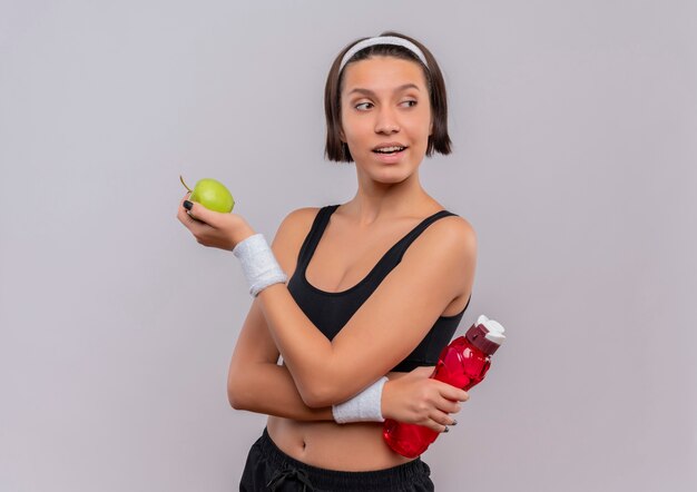 Mulher jovem fitness em roupas esportivas segurando uma maçã verde e uma garrafa de água olhando para o lado com um sorriso confiante em pé sobre uma parede branca