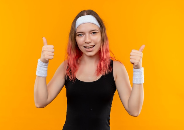 Mulher jovem fitness em roupas esportivas sorrindo alegremente mostrando os polegares para cima com as duas mãos em pé sobre a parede laranja