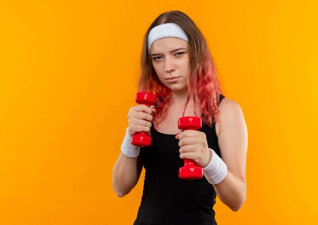 Mulher jovem fitness em roupas esportivas usando halteres, fazendo exercícios, parecendo confiante em pé sobre a parede laranja