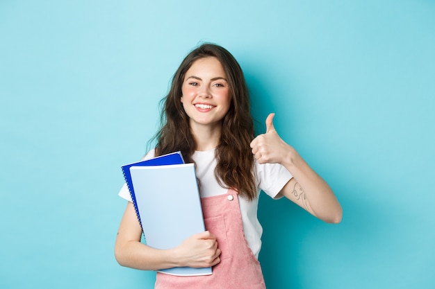 Mulher jovem frequenta cursos, aluna estudando, segurando cadernos e mostrando o polegar em aprovação, recomendando a empresa, em pé sobre um fundo azul