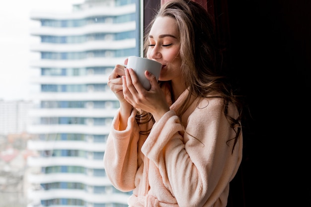 Foto grátis mulher jovem fresca em um roupão rosa beber chá, olhando pela janela.