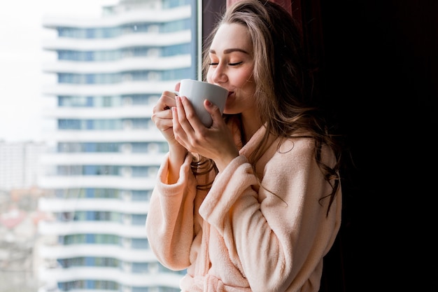 Mulher jovem fresca em um roupão rosa beber chá, olhando pela janela.