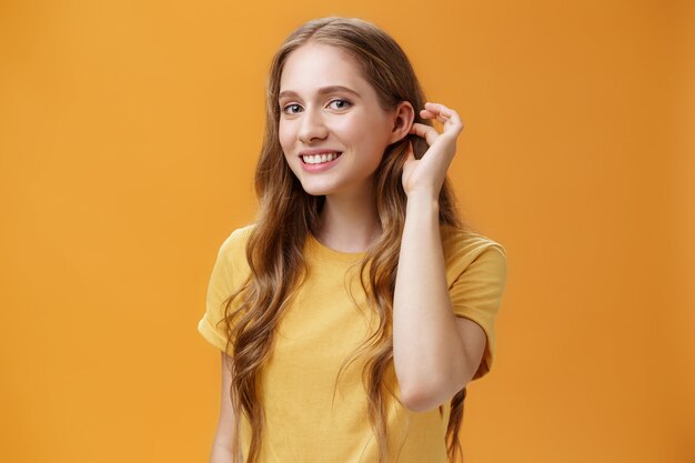 Mulher jovem glamour bonita e terna com penteado ondulado fofo sacudindo a mecha de cabelo atrás da orelha, olhando para a câmera com tímido sorriso sensual, olhando com admiração para o cara sobre fundo laranja. Copie o espaço