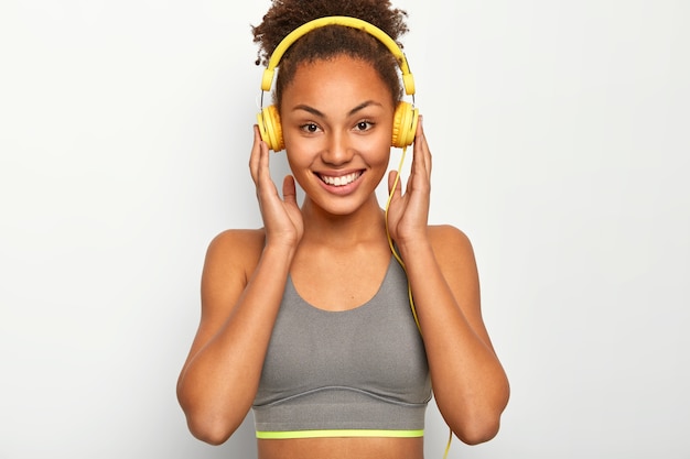 Mulher jovem gosta de música como motivação pessoal, mantém as duas mãos nos fones de ouvido, sorri agradavelmente e usa sutiã esportivo cinza
