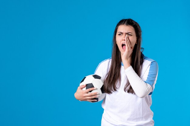 Mulher jovem gritando de frente segurando uma bola de futebol no azul