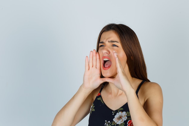 Mulher jovem gritando, segurando as mãos perto da boca em um top floral e parecendo descontente