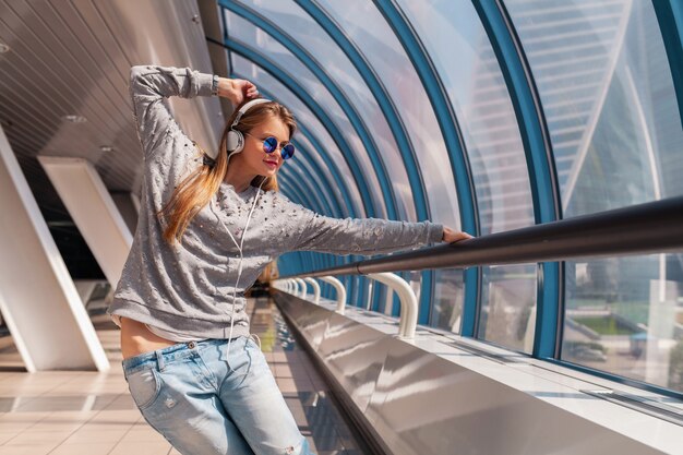 Mulher jovem hippie dançando se divertindo em um edifício urbano moderno, vestida com roupa casual e ouvindo música em fones de ouvido