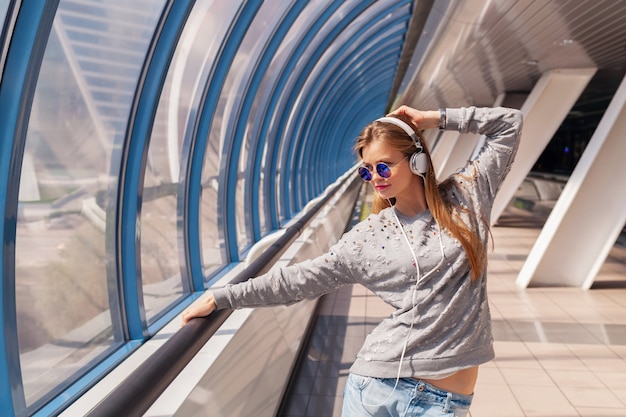 Mulher jovem hippie em roupa casual se divertindo ouvindo música em fones de ouvido, vestindo jeans, suéter e óculos escuros, estilo urbano
