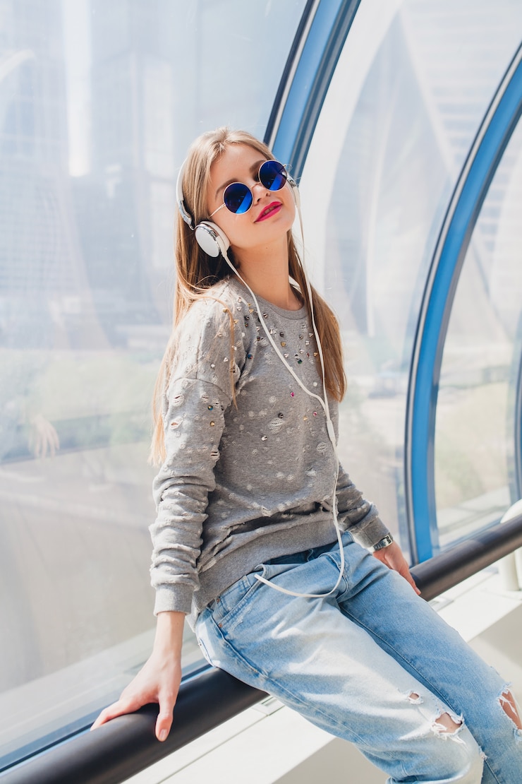 Foto grátis mulher jovem hippie em roupa casual se divertindo ouvindo música em fones de ouvido, vestindo jeans, suéter e óculos escuros, estilo urbano