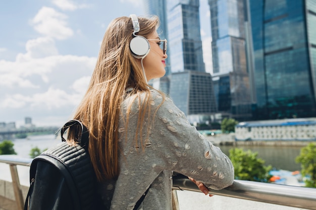 Mulher jovem hippie se divertindo na rua ouvindo música em fones de ouvido, usando óculos escuros cor de rosa e mochila, estilo urbano primavera verão