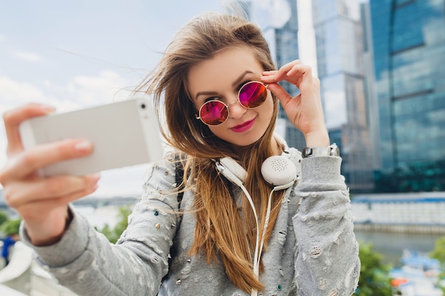 Mulher jovem hippie se divertindo na rua, usando óculos escuros rosa, estilo urbano primavera verão, tirando selfie pisture no smartphone