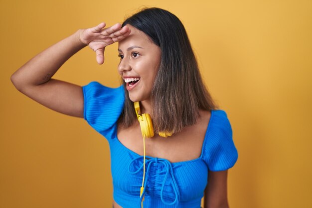 Mulher jovem hispânica em pé sobre fundo amarelo muito feliz e sorrindo olhando para longe com a mão sobre a cabeça. conceito de pesquisa.